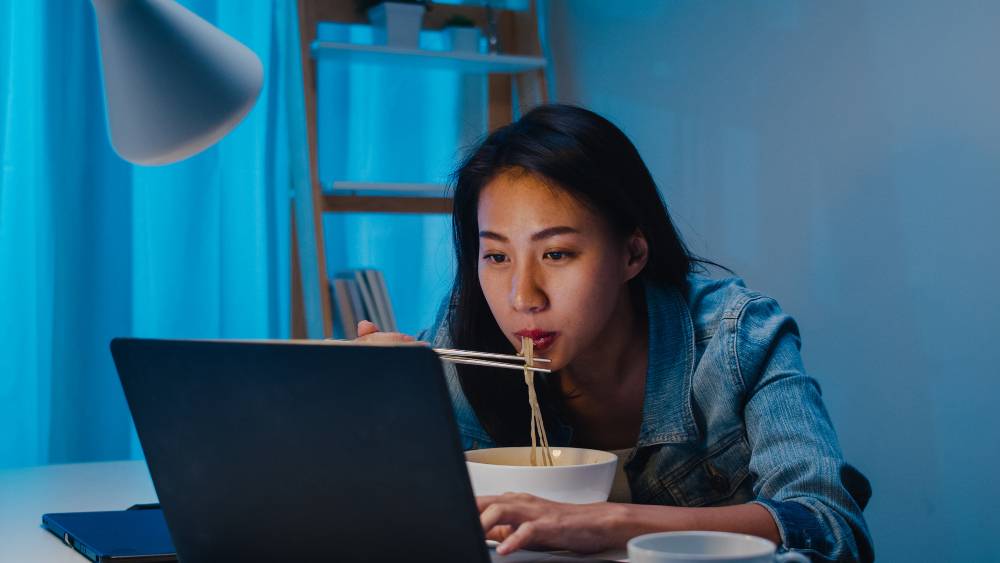 ilustrasi perempuan membuka laptop dan makan mie instan saat begadang (freepik)